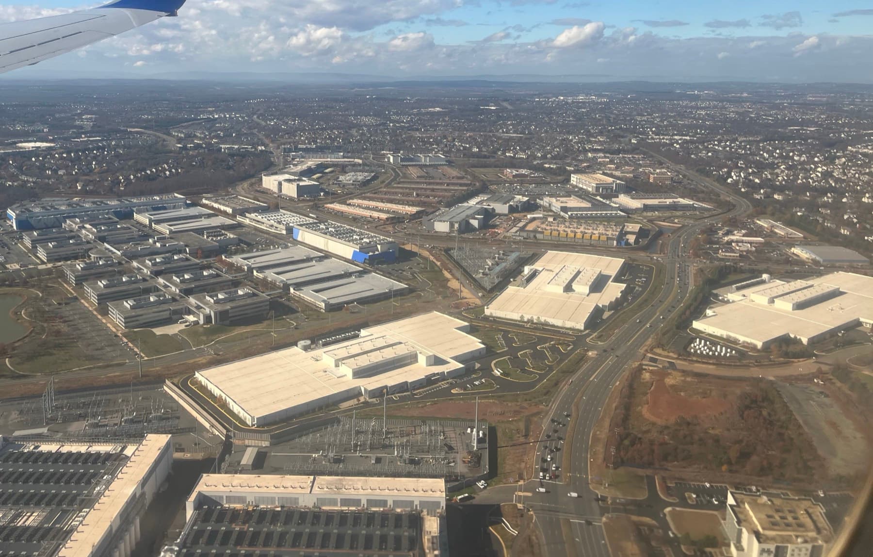 Aerial view of a dense cluster of long, low, windowless data center buildings surrounded by parking lots, electrical yards and roads, with tree cover and suburban development at the edges.