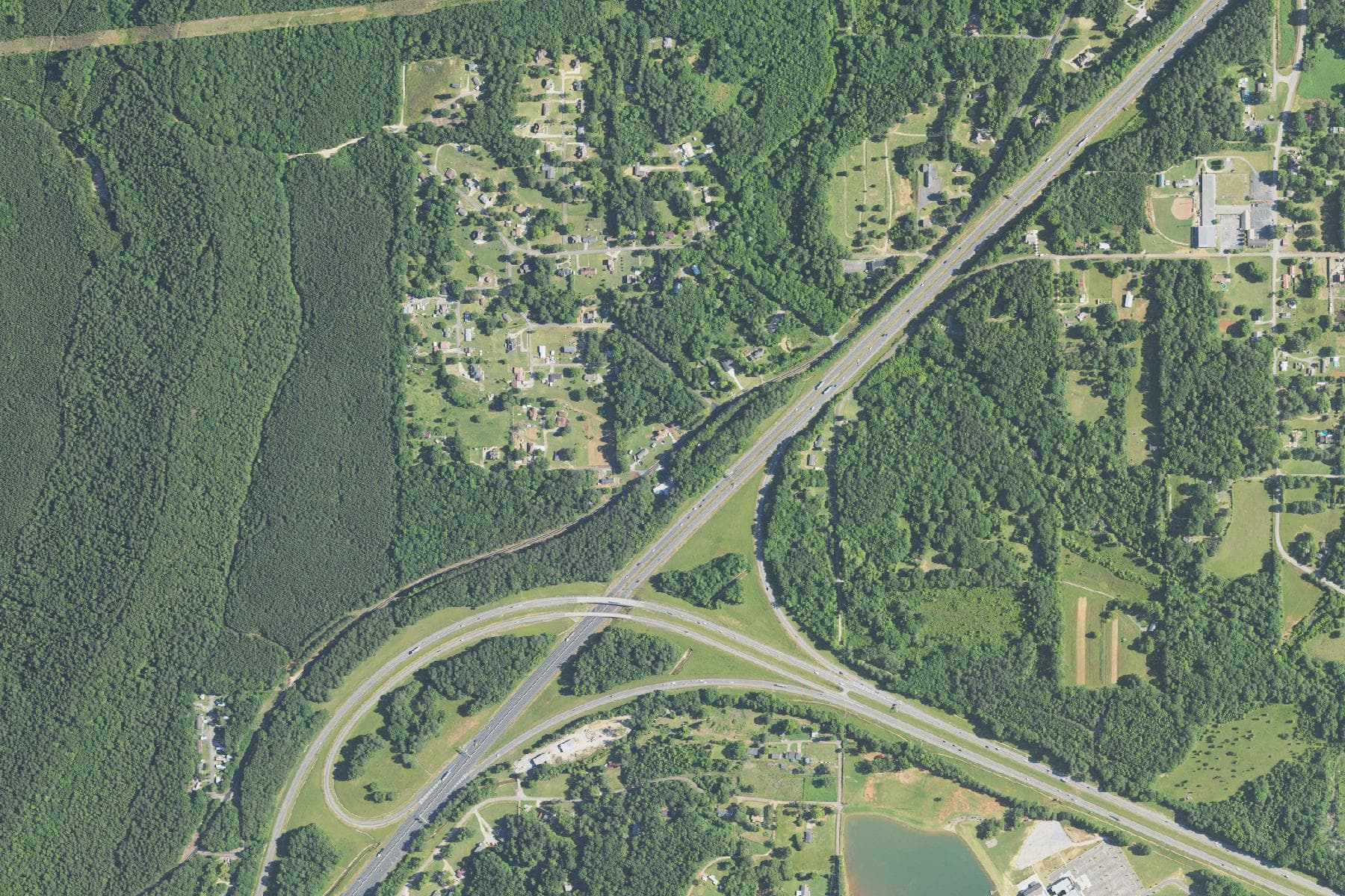 Overhead view of a large unbroken tract of dense hardwood forest on the western side of a major highway interchange, with scattered houses and yards along the roads to the east.