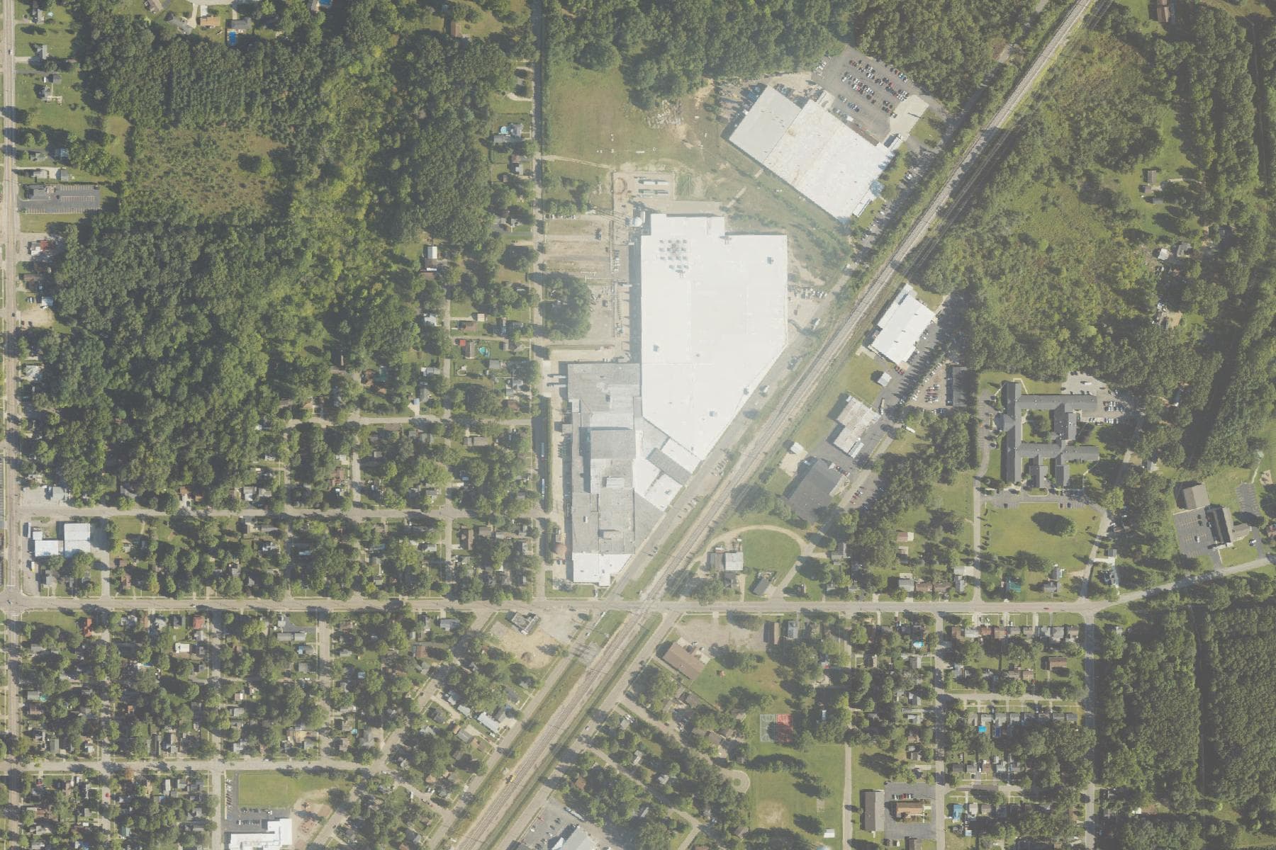 Overhead view of a large white-roofed industrial plant beside a rail line in a small town, with streets of single-family houses immediately to the west and south.