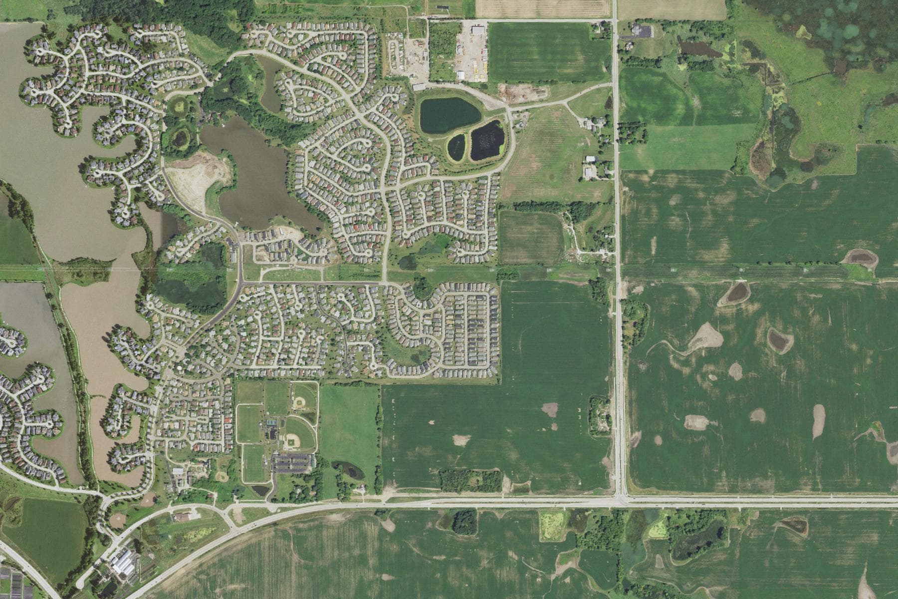 Overhead view of a dense curving subdivision around lakes on the west side of a straight north-south road, with open farm fields stretching east of the road.