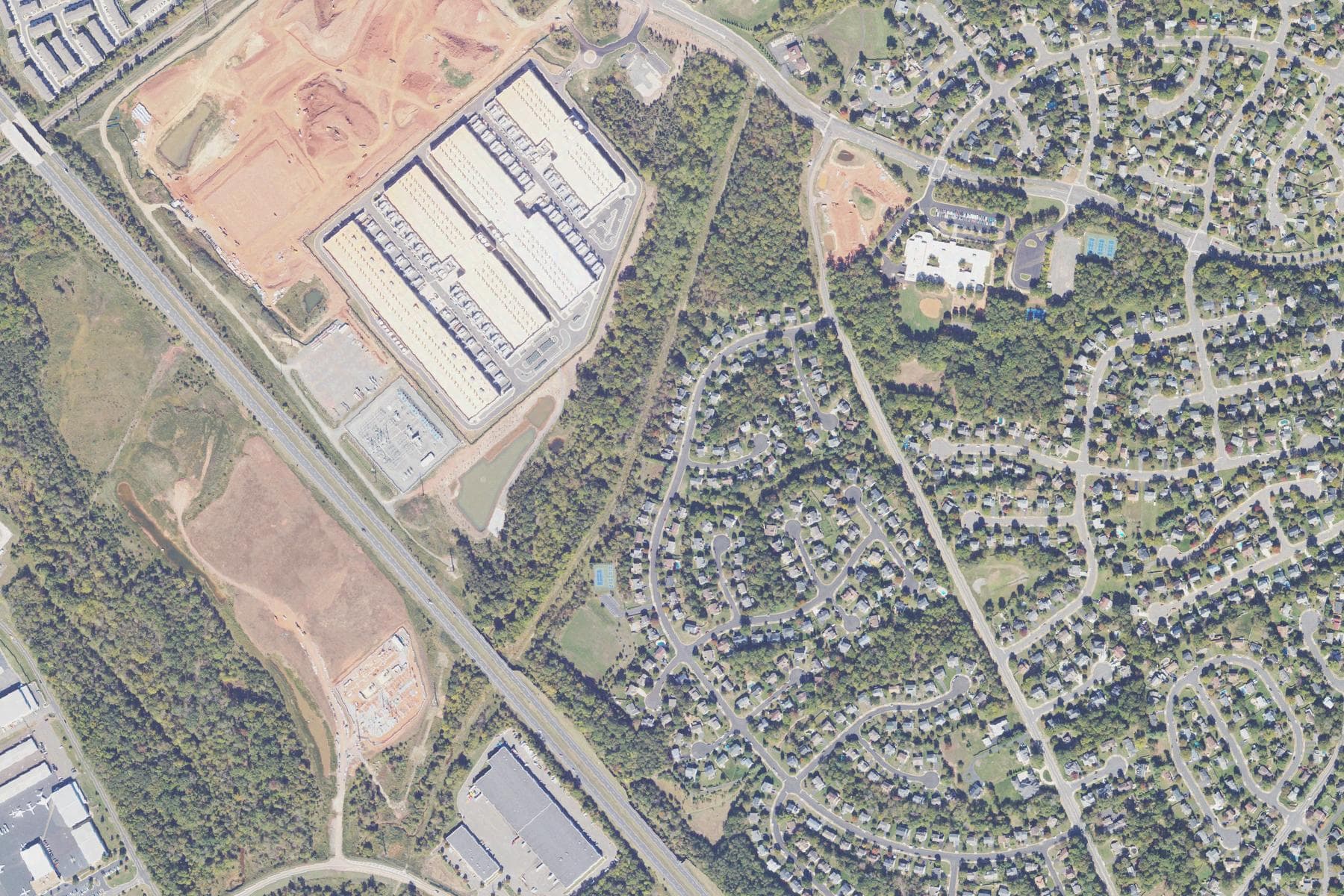 Overhead view of four very large windowless buildings with rooftop cooling units and an adjacent electrical substation, separated by a narrow band of trees from a dense subdivision of single-family houses with back gardens and swimming pools.
