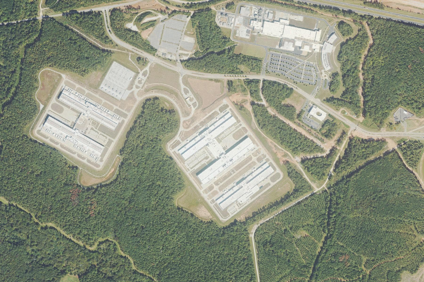Overhead view of two clusters of long windowless data center buildings on cleared pads cut into dense forest, with a large manufacturing plant and its parking lots to the north beside an interstate.
