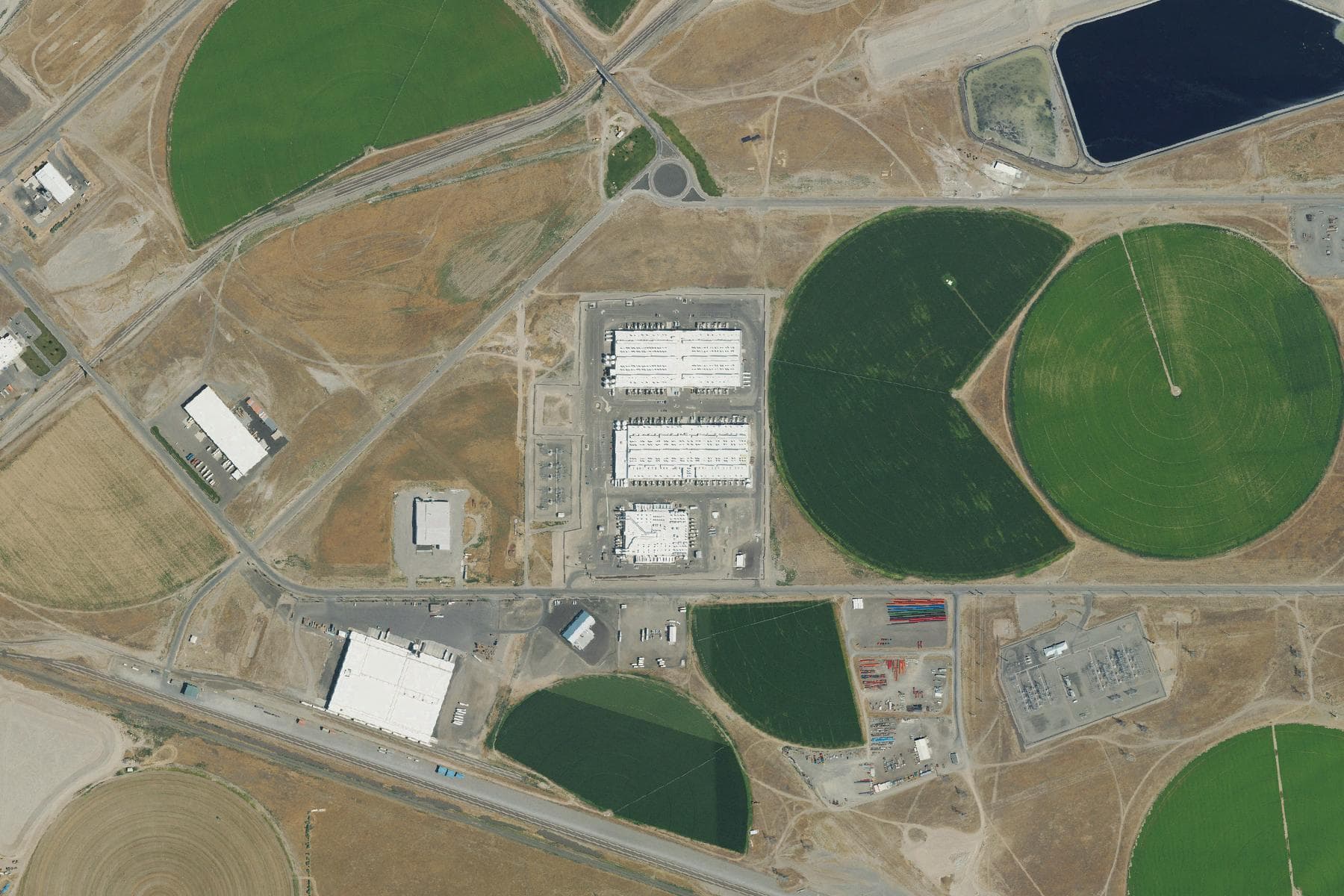 Overhead view of three large windowless data center buildings with rows of rooftop cooling units, ringed by bright green circular irrigation fields, a fenced electrical substation to the southeast and a dark rectangular lagoon to the northeast.