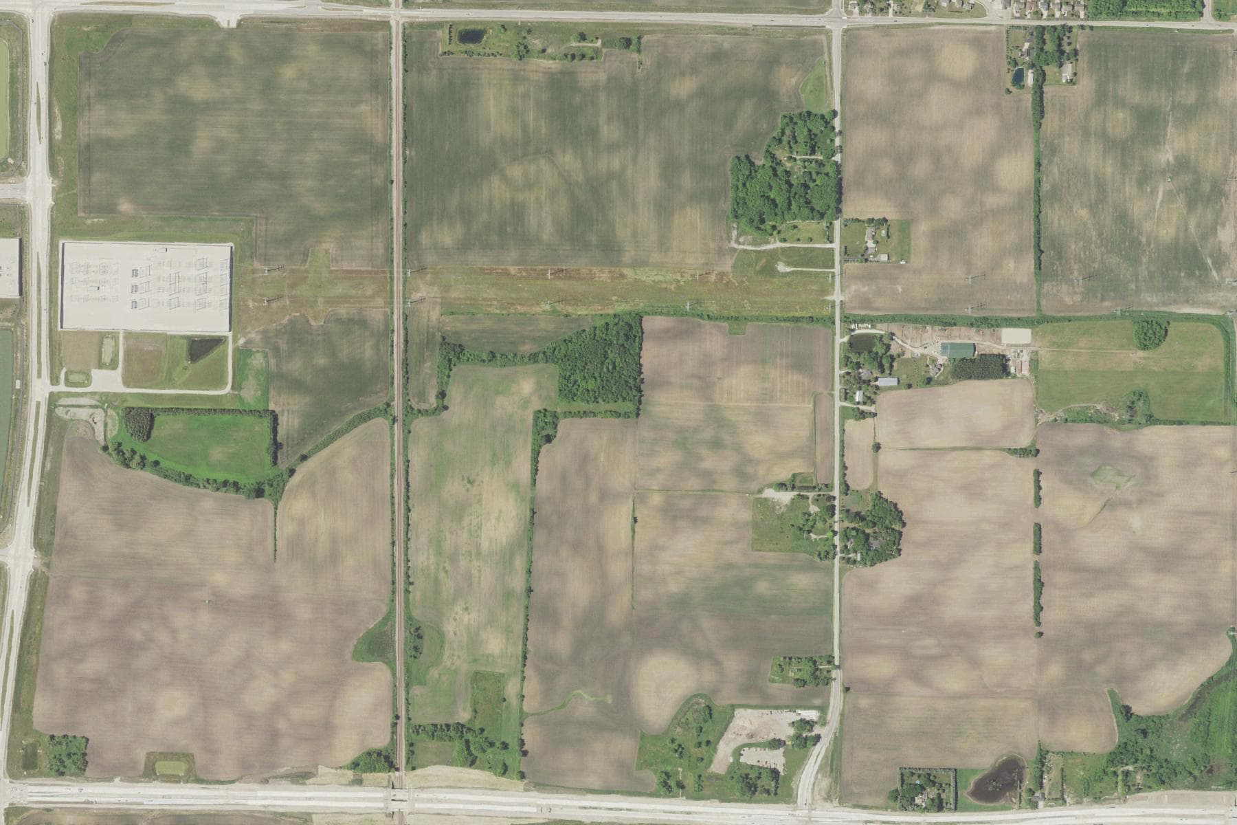 Overhead view of flat open farm fields divided by section roads and a rail line, with a single large industrial building at the western edge and scattered farmhouses.