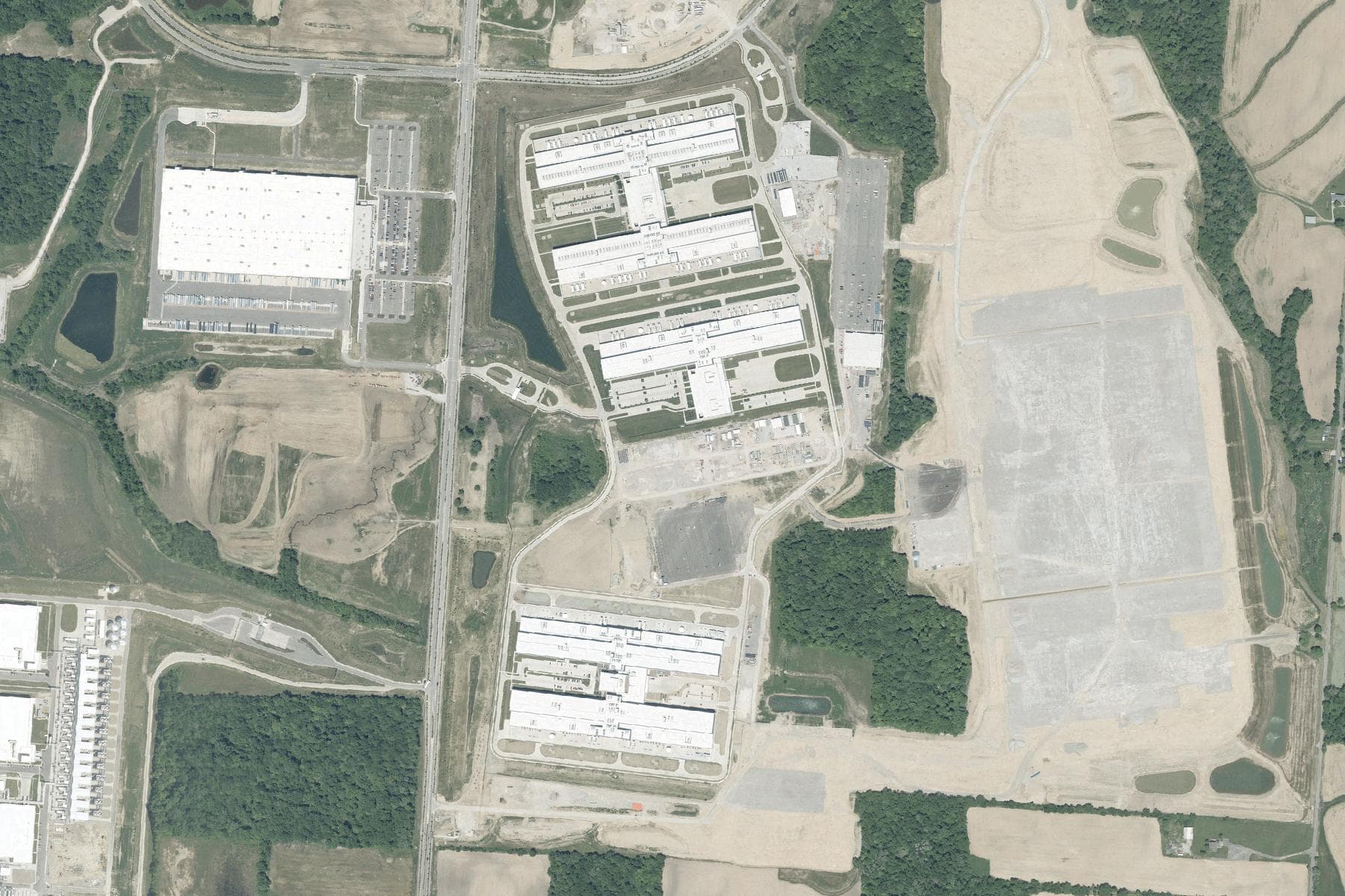 Overhead view of several very large windowless buildings arranged in rows, with a vast graded earth pad of comparable size cleared to the east, surrounded by woodland and farm fields.