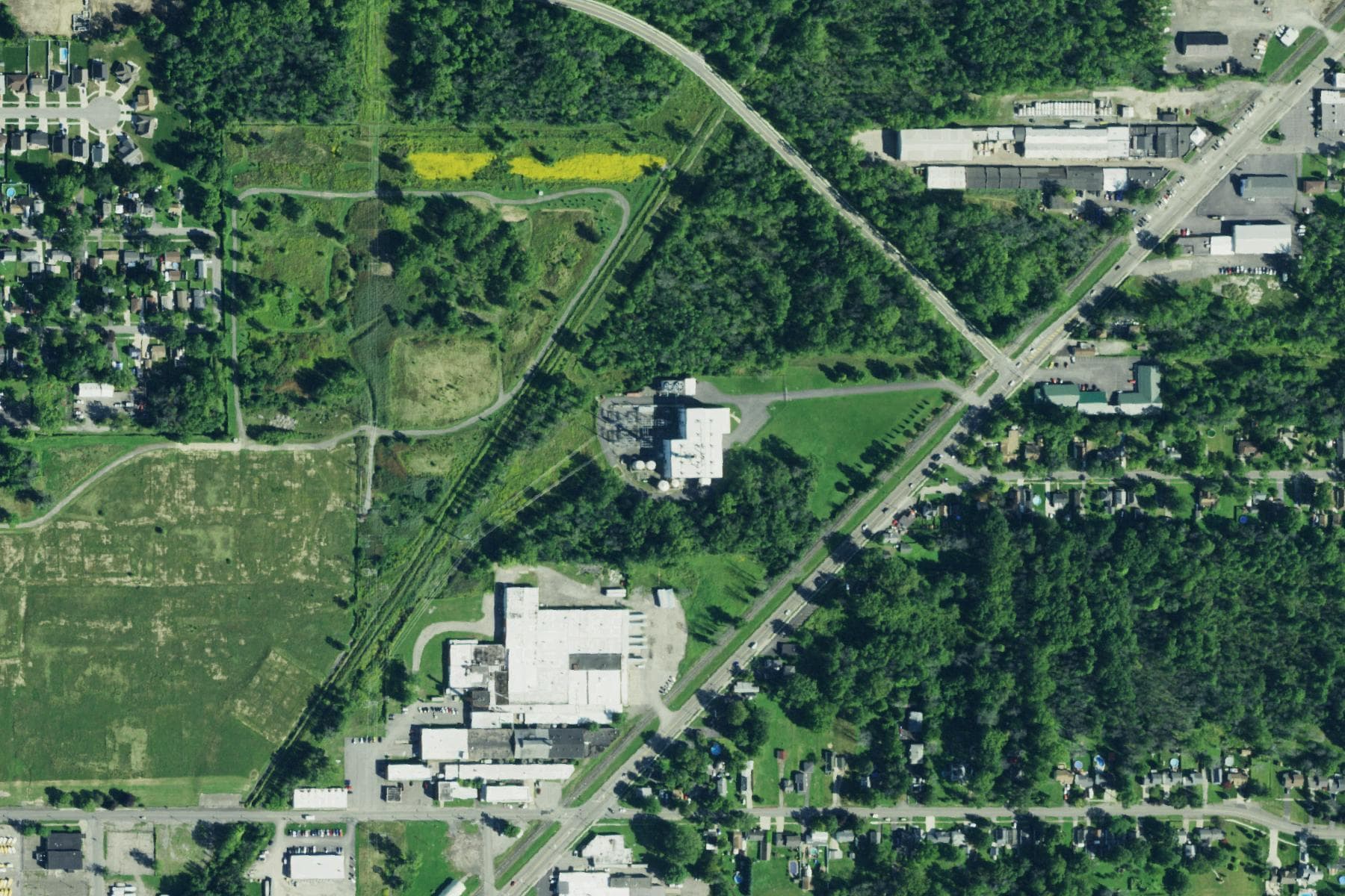 Overhead view of a small gas-fired power station with storage tanks and a switchyard on a wooded parcel, with residential streets on three sides and a rail line and highway to the south.