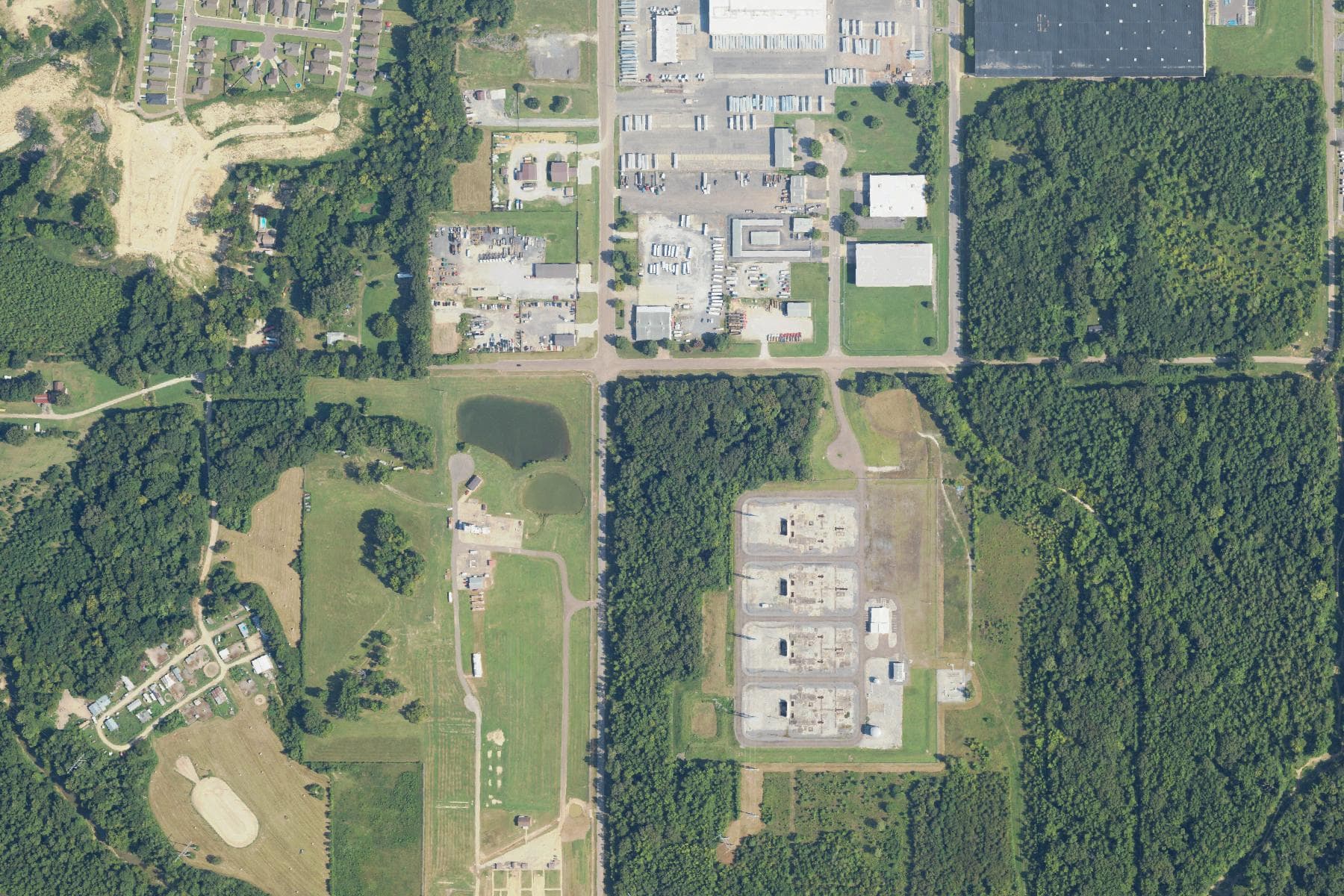 Overhead view of a fenced industrial compound holding four large rectangular concrete foundation pads in a clearing, surrounded by woodland, with houses and a trucking yard a short distance to the north and west.