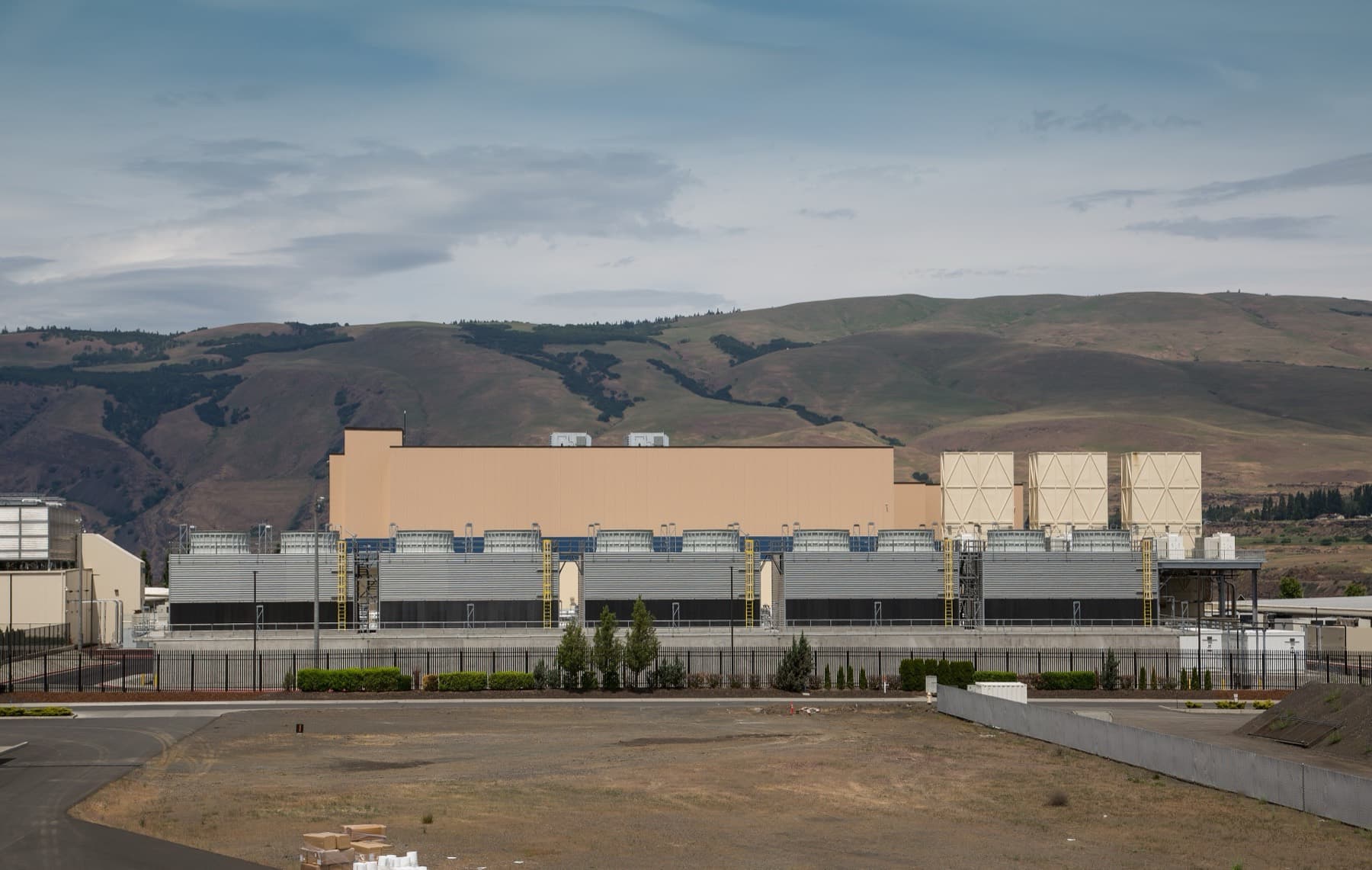 A low industrial building with cooling equipment on its roof beside a river valley, with dry hills rising behind it.
