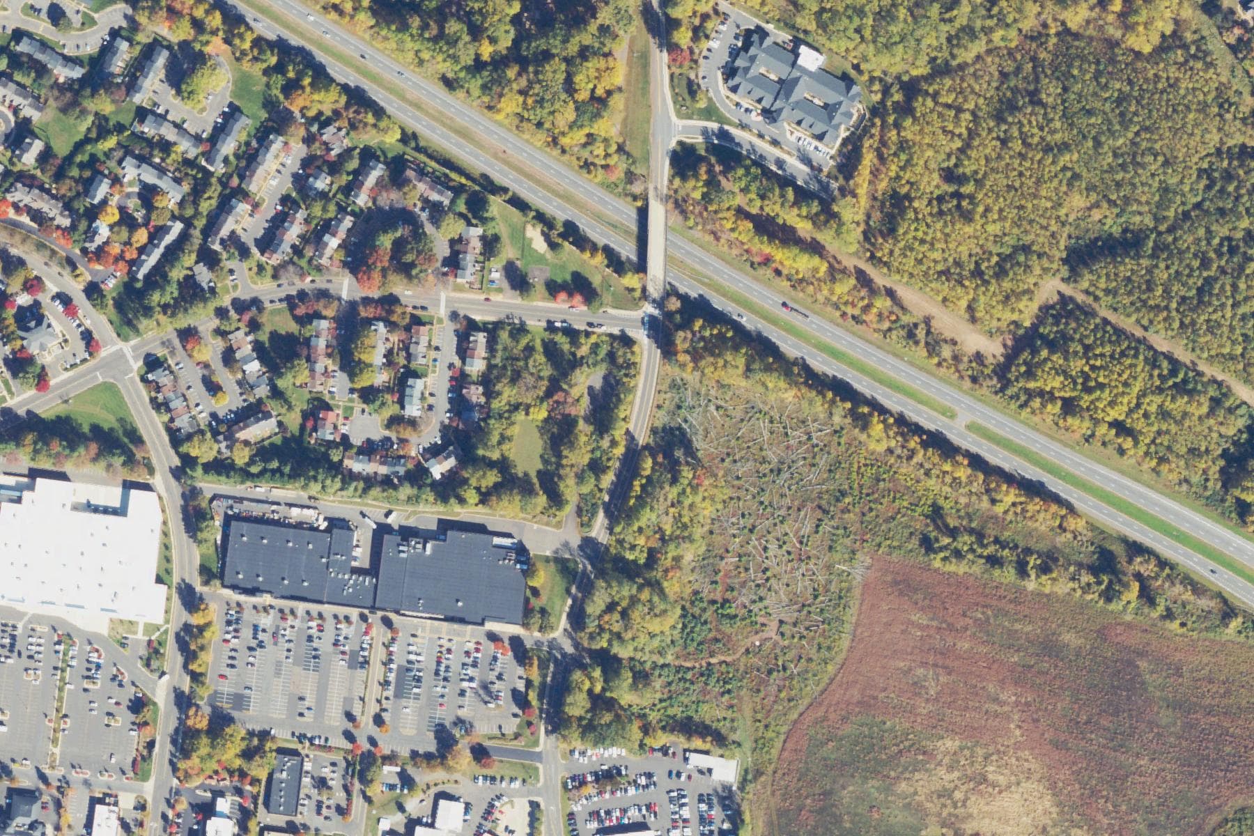 Overhead view of a cleared parcel of open ground at a highway junction on the edge of a small town, bordered by trees, commercial buildings and residential streets.