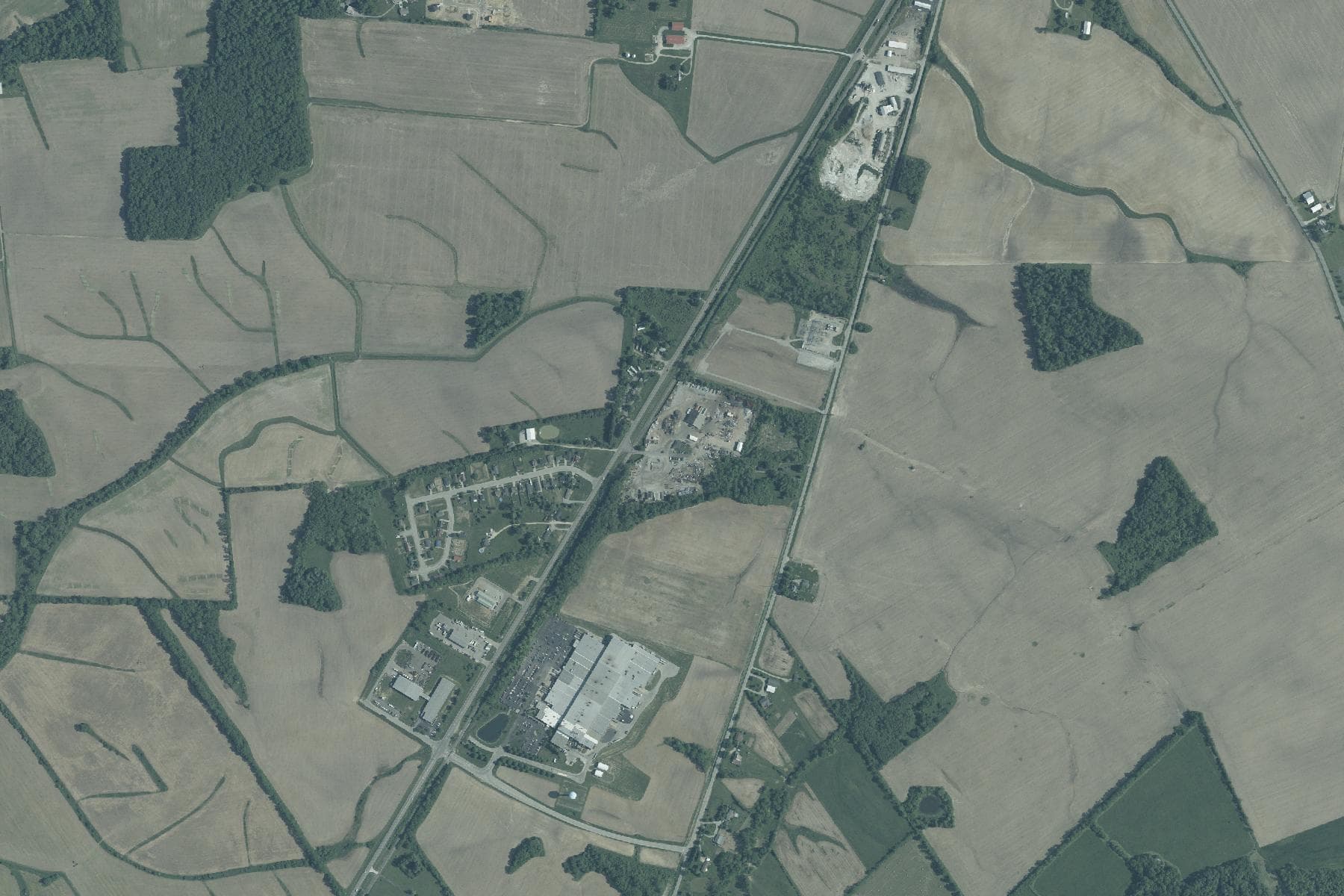 Overhead view of flat farm fields and woodlots along a two-lane highway on the edge of a small city, with scattered houses and farm buildings.