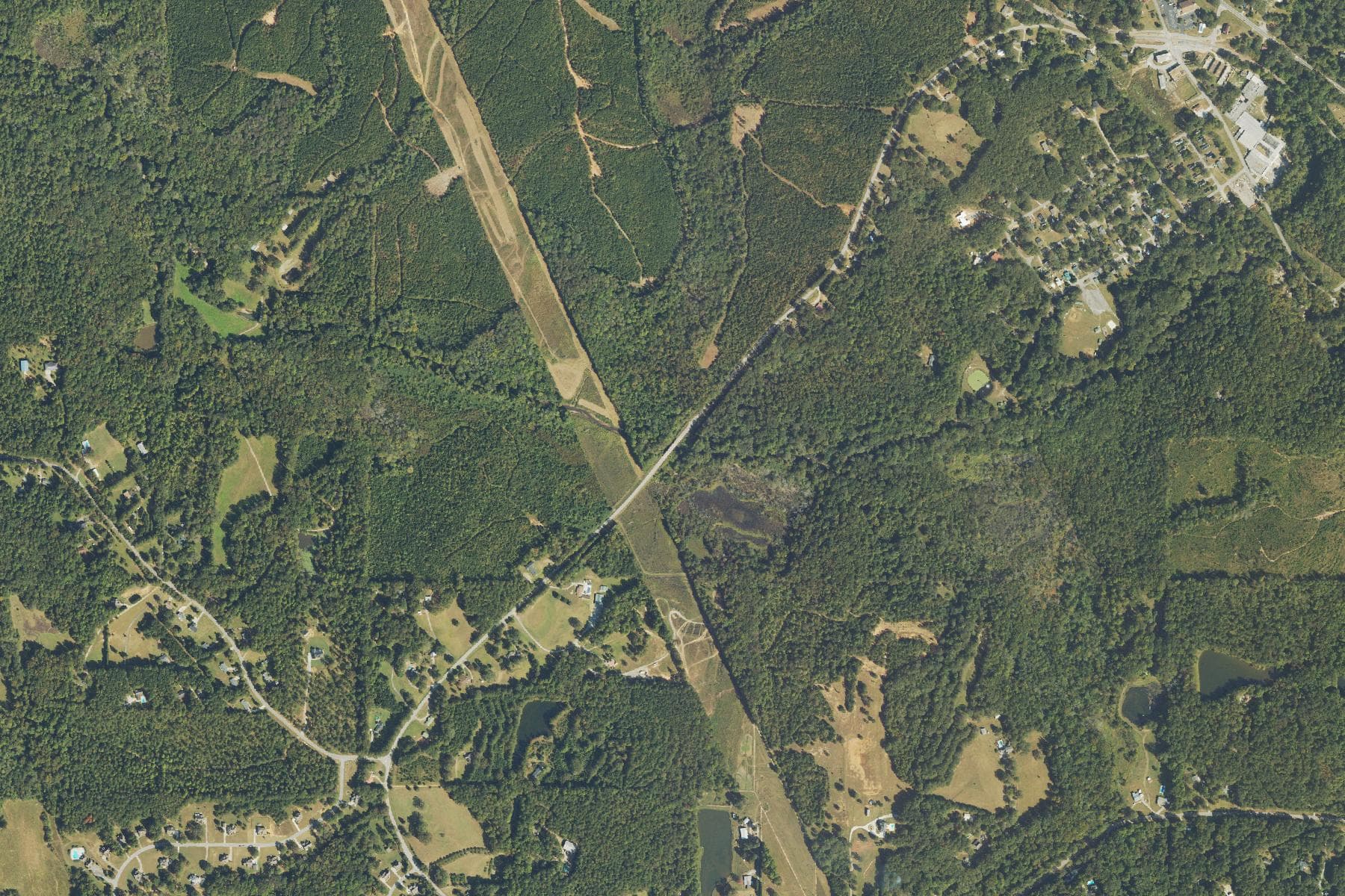 Overhead view of rolling pine and hardwood forest crossed by a straight cleared utility corridor and a winding rural road, with scattered houses and small pastures and a small town at the upper right.