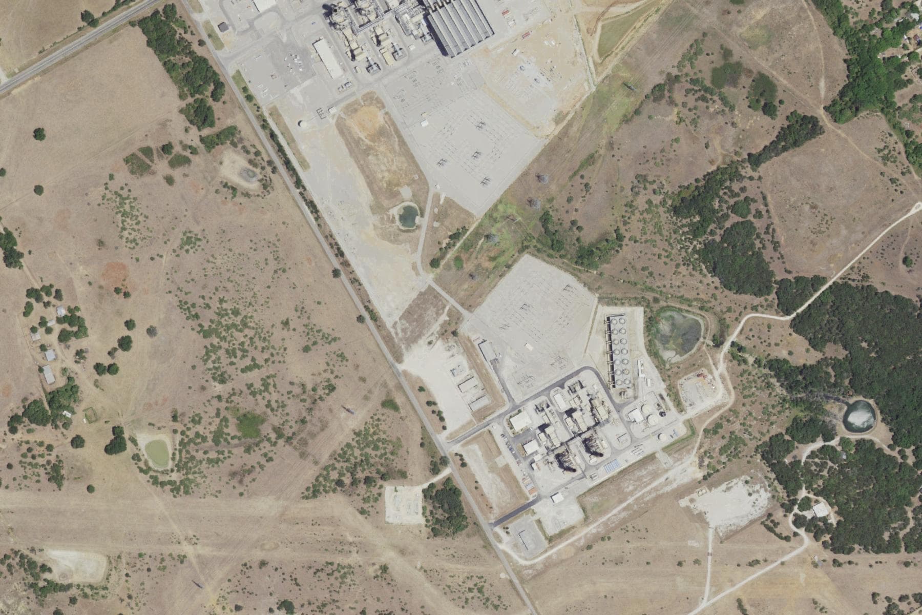 Overhead view of a gas-fired generating plant and a second fenced industrial yard on pale open ground, surrounded by dry ranchland, stock ponds, scattered houses and small holdings.