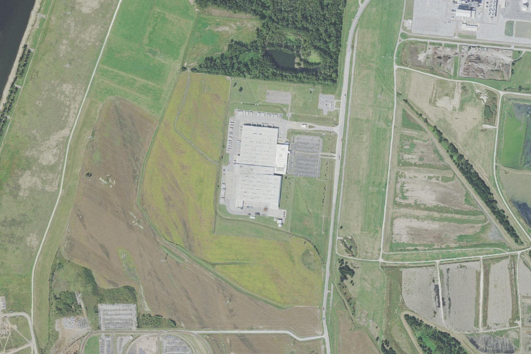 Overhead view of a very large rectangular factory building with extensive parking and loading areas, set among woodland and low-density residential streets in an industrial district.