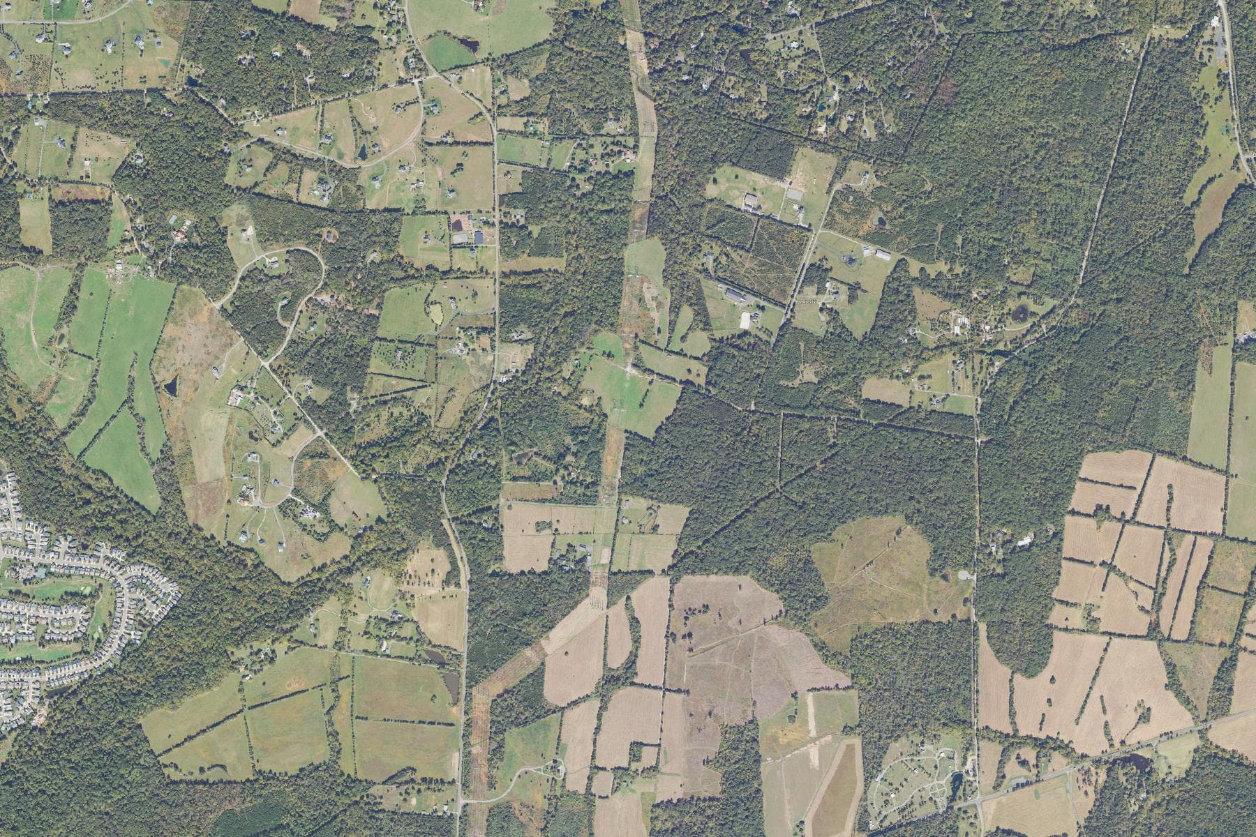 Overhead view of a broad corridor of open farm fields, pasture and woodlots along a rural two-lane road, bordered by protected parkland to the south.