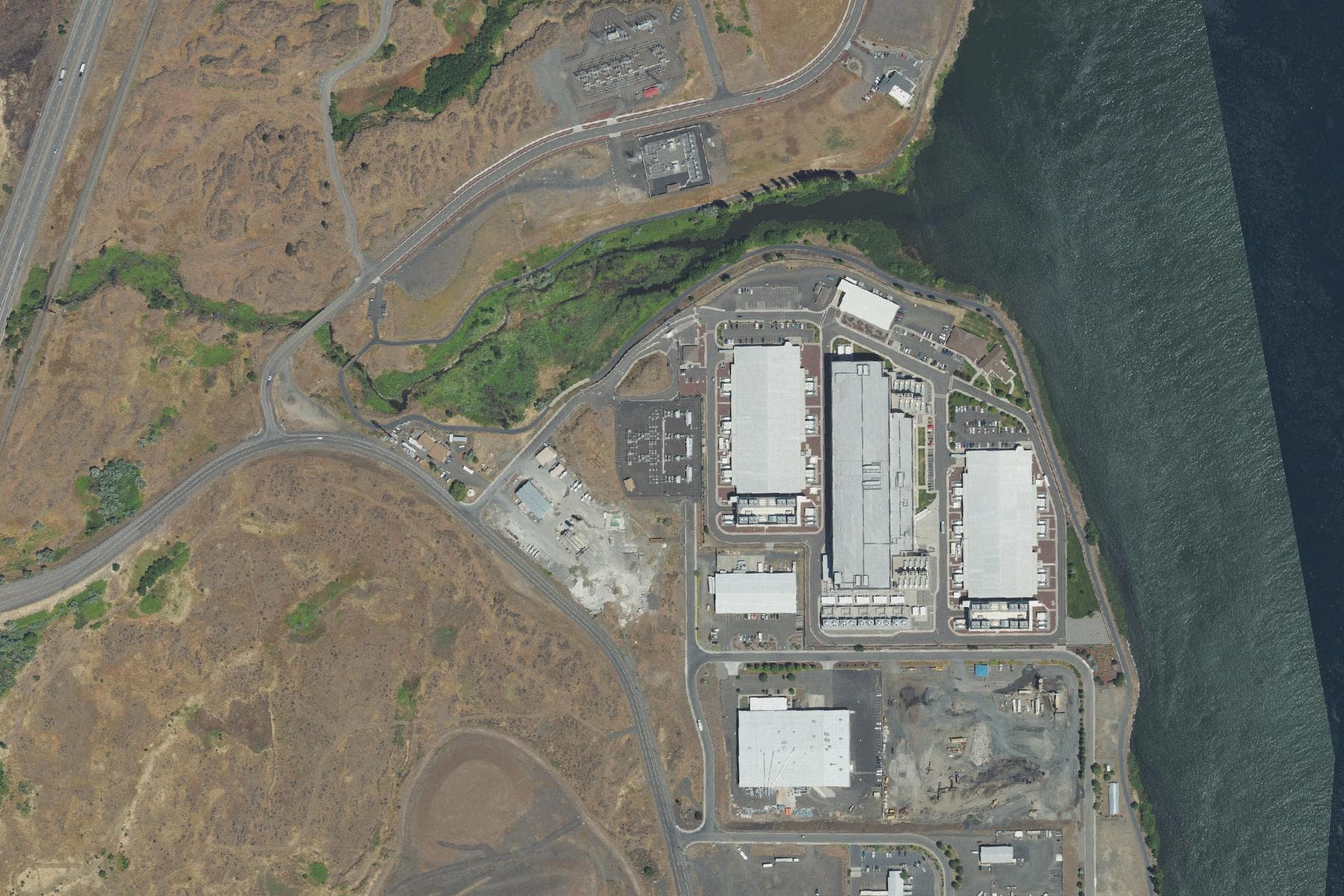 Overhead view of three long data center halls with rows of cooling units along their flanks, an electrical switchyard to the north, and a wide river running along the eastern edge of the site.