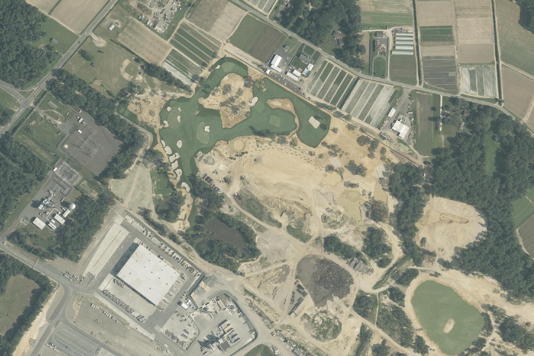 Overhead view of an industrial parcel with large flat-roofed buildings and equipment yards beside a two-lane road, with houses along the road and woodland behind.